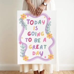 May include: A white poster with the text "TODAY IS GOING TO BE A GREAT DAY" in colorful bubble letters. The text is framed by a wavy purple border, flowers, leaves, and starbursts. The poster is held by a person.