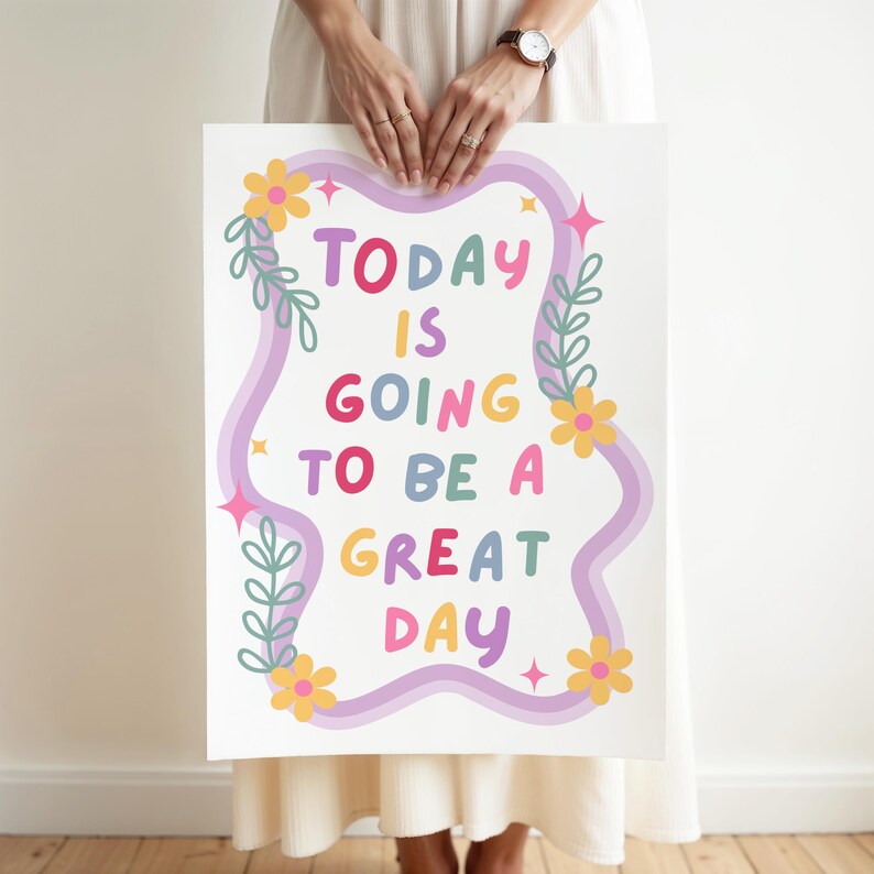May include: A white poster with the text "TODAY IS GOING TO BE A GREAT DAY" in colorful bubble letters. The text is framed by a wavy purple border, flowers, leaves, and starbursts. The poster is held by a person.