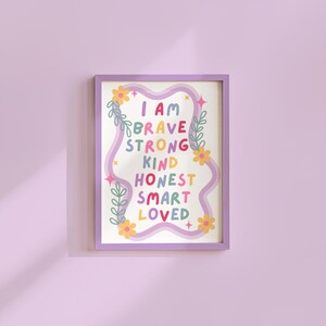 May include: A framed art print with a purple border. The print features the words "I AM BRAVE STRONG KIND HONEST SMART LOVED" in colorful, playful lettering. Floral and leaf accents surround the text on a white background.