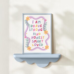 May include: A framed art print with the words "I AM BRAVE STRONG KIND HONEST SMART LOVED" in colorful letters. The print is decorated with floral and wavy design elements in a light blue frame, resting on a matching shelf.