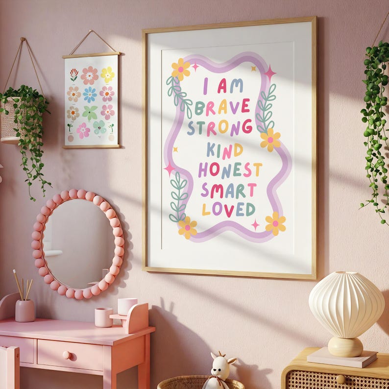 May include: A framed art print with the words "I AM BRAVE STRONG KIND HONEST SMART LOVED" in colorful text. The print is surrounded by a decorative border with flowers and leaves. A pink vanity with a round mirror and a floral print are also visible.