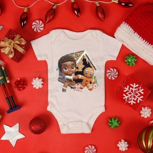 May include: A white baby onesie with a gingerbread house, a gingerbread man, and a cartoon boy with brown skin. The boy is holding the gingerbread man's hand.
