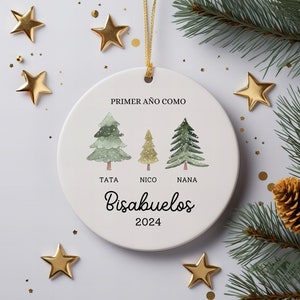 May include: White ceramic Christmas ornament with a gold string, featuring watercolor illustrations of three Christmas trees and the text "PRIMER AÑO COMO Bisabuelos 2024". Gold star decorations and pine branches surround the ornament.