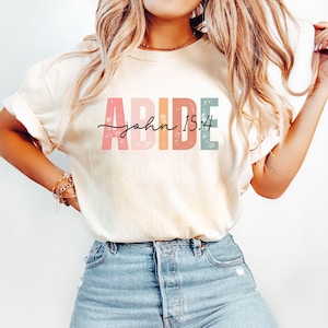 May include: A white t-shirt with the word "ABIDE" in colorful letters and the text "john 15:4" in black script.