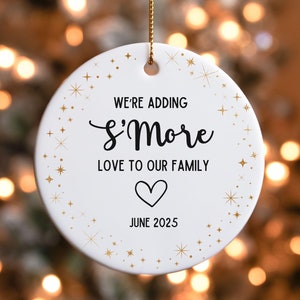 May include: White round ceramic ornament with gold glitter stars and the text "We're adding S'more love to our family June 2025" with a heart symbol.