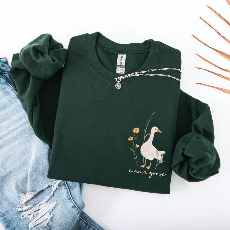 Pu&ograve; includere: Una felpa verde scuro con un'oca bianca e fiori gialli stampati sul davanti. Il testo "mama goose" &egrave; stampato sotto l'oca.