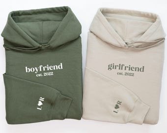 Puede incluir: Dos sudaderas con capucha, una verde y otra beige, con las palabras "boyfriend est. 2022" y "girlfriend est. 2022" impresas en ellas. Cada sudadera tiene un pequeño corazón en la manga.