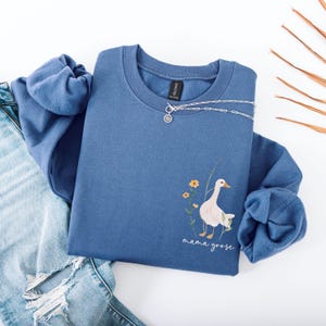 Pu&ograve; includere: Una felpa blu con un'oca bianca e fiori stampati sul davanti. Il testo "mama goose" &egrave; stampato sotto l'oca.