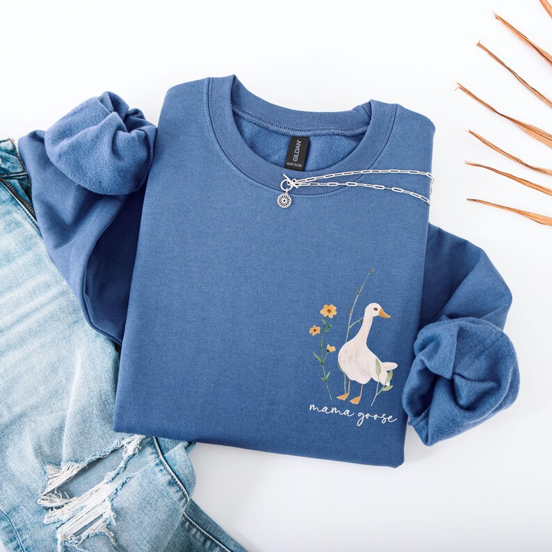 Pu&ograve; includere: Una felpa blu con un'oca bianca e fiori stampati sul davanti. Il testo "mama goose" &egrave; stampato sotto l'oca.