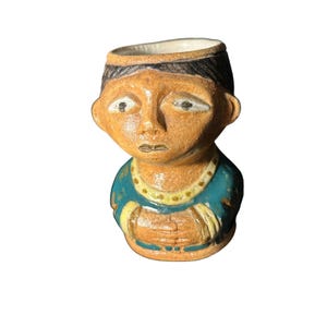 Pode incluir: Estatueta de cerâmica em forma de cabeça e parte superior do corpo de uma pessoa. A estatueta tem um rosto bronzeado, cabelo preto e uma camisa azul-petróleo. A parte superior é oca, possivelmente para um pequeno vaso ou recipiente.