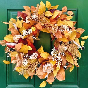 May include: A vibrant autumn wreath with a mix of colorful leaves in shades of yellow, orange, and red. The wreath is adorned with white floral accents and faux berries, creating a festive and welcoming seasonal decoration.