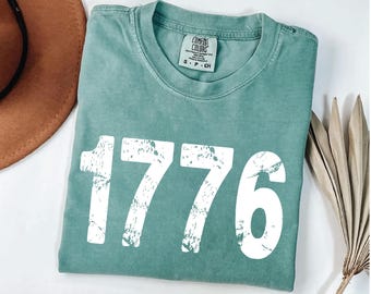 May include: A teal-colored t-shirt with the number "1776" printed in distressed white lettering. The shirt has a crew neck and short sleeves. The tag reads "Comfort Colors".