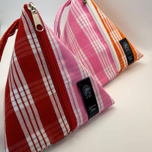 May include: Two triangular zippered pouches. One is red with white stripes, the other is pink with white stripes and orange accents. Each has a zipper closure and a small fabric tag with text. The pouches are likely for cosmetics or small items.
