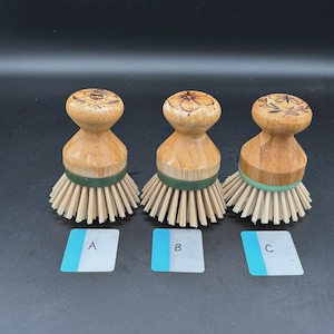 May include: Three wooden dish scrub brushes with engraved floral designs on the tops. Each brush has a light brown handle, tan bristles, and a green or teal band. Small white and blue labels are below each brush.