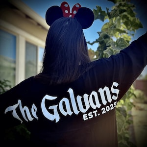 May include: Black long-sleeve shirt with the text "The Galvans EST. 2025" in white script. The person is wearing a Minnie Mouse ear headband with black ears and a red polka dot bow.