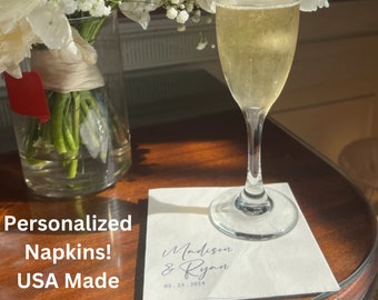 May include: Personalized napkins with the text "Madison & Ryan" and the date "05.25.2024" printed on them. The napkins are white and are sitting on a wooden table next to a glass of champagne.
