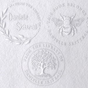 May include: Three circular embossed designs on white paper. The top left features a leaf wreath with the text "From the library of Danielle Skittrall". The top right shows a bee with "This book belongs to Danielle Skittrall". The bottom shows a tree.