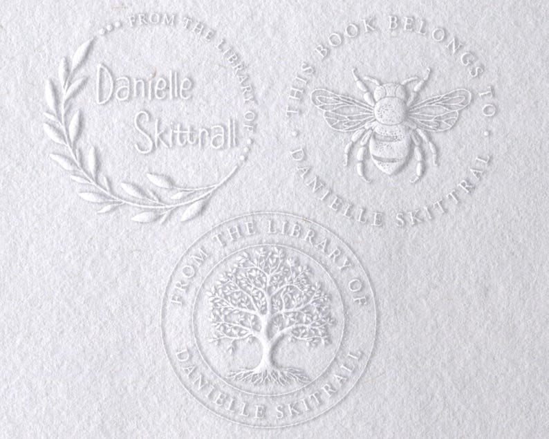 May include: Three circular embossed designs on white paper. The top left features a leaf wreath with the text "From the library of Danielle Skittrall". The top right shows a bee with "This book belongs to Danielle Skittrall". The bottom shows a tree.