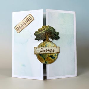 May include: A white gatefold card featuring a light blue marbled background. The left panel displays a rectangular tag with the word IMAGINE. The center shows a globe topped with a green tree and a banner across the middle with the word Dreams.