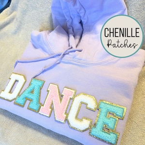 May include: A lavender hoodie with the word "DANCE" spelled out in colorful chenille patches. The patches are in white, pink, light blue, and gold. The hoodie has a drawstring hood and is made of a soft, comfortable fabric.