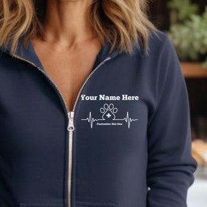 May include: Navy blue zip-up hoodie with white text that reads "Your Name Here" above a paw print design with a cross inside, and the text "Customize this line" below. The zipper is silver.