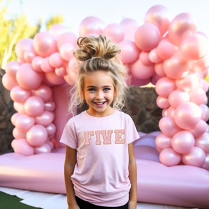 Peut inclure: Une jeune fille aux cheveux blonds en chignon désordonné sourit à la caméra. Elle porte un t-shirt rose avec le mot "FIVE" en lettres dorées pailletées. La fille est debout devant une arche de ballons roses.