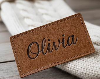 May include: A rectangular, brown leather patch with the name "Olivia" engraved in a cursive font. The patch has a stitched border and is set against a white, cable-knit background.