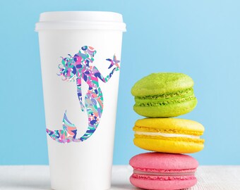May include: A white paper coffee cup with a colorful mermaid design holding a starfish. Three stacked macarons in green, yellow, and pink sit to the right of the cup. The background is a light blue.