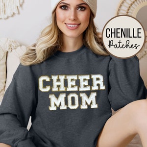 May include: A dark gray sweatshirt with the words "CHEER MOM" in gold chenille lettering. The sweatshirt is a crew neck style and has long sleeves.