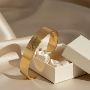 May include: A gold-colored cuff bracelet with the name "Brianna" engraved in a cursive font. The bracelet is displayed inside an open, off-white gift box with a satin lining. The background features a soft, draped fabric.