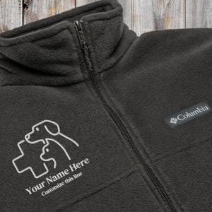 May include: A dark grey fleece jacket with a zip fastening. The jacket features a white embroidered logo of two dogs and a cross with the text "Your Name Here Customize this line" underneath. The jacket also has a black Columbia logo patch on the right sleeve.
