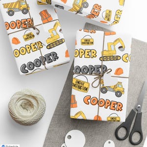May include: Gift-wrapped presents with construction-themed wrapping paper. The paper features yellow construction vehicles, orange hard hats, and the name "COOPER" in bold letters. Each gift is tied with twine and a tag.
