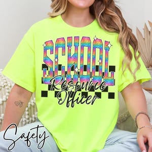 May include: A bright yellow t-shirt featuring the text "SCHOOL Resource Officer" in a rainbow gradient. The design incorporates black chequered squares and star shapes. The word "Safety" is written in cursive script at the bottom.
