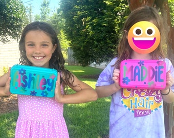 May include: Two rectangular plastic pencil cases. One is light blue with the name Ashley printed in dark blue letters and decorated with small blue flowers. The other is bright pink with the name Maddie printed in dark green letters.