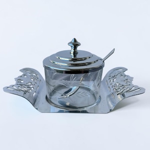 May include: A clear glass condiment jar with a silver-toned metal lid and a matching silver-toned metal spoon. The jar features etched leaf patterns and sits within a decorative silver-toned metal stand with ornate, wing-like side handles.