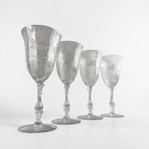 May include: A set of four clear glass goblets arranged in a row, each featuring an intricate etched floral pattern on the bowl and a decorative, textured stem. The glasses vary in size, with the largest positioned in the front and the smallest in the back.