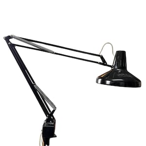 May include: A black articulated desk lamp with a conical shade. The lamp features a spring-loaded arm and a pivoting head, allowing for adjustable positioning. The lamp is designed for focused lighting and is suitable for a desk or workspace.