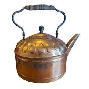 May include: A vintage copper tea kettle with a dark wooden handle and a curved metal handle. The kettle has a decorative pattern on the lid and a spout for pouring. The copper has a warm, aged patina.