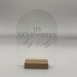 Peut inclure: Panneau en acrylique transparent avec un sommet en forme d'arche, portant l'inscription "LES GOURMANDISES des Kids" en lettres grises. Le panneau est maintenu par une petite base en bois rectangulaire. Le fond est blanc.