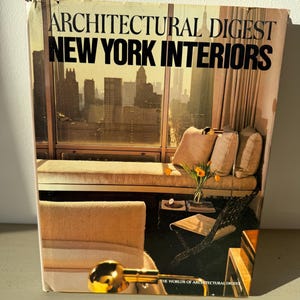 Może przedstawiać: Okładka magazynu Architectural Digest New York Interiors. Na okładce znajduje się zdjęcie pokoju z oknem z widokiem na panoramę miasta. Tekst "ARCHITECTURAL DIGEST NEW YORK INTERIORS" jest wyświetlany u góry.