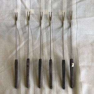 May include: Set of six silver fondue forks with dark brown wooden handles. The forks have three prongs and are arranged in a row.