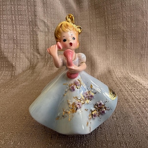 May include: A porcelain figurine of a girl in a blue dress with gold and purple floral accents. She is holding a pink rotary phone to her ear and has a surprised expression on her face.