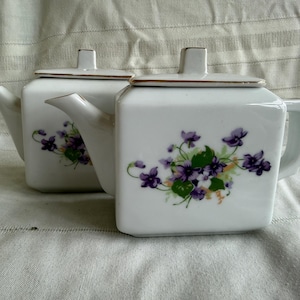 May include: Two white porcelain teapots with square bodies and matching lids. Each teapot features a floral design of purple violets and green leaves. The rims and lid edges have a gold trim. The teapots are displayed on a white surface.