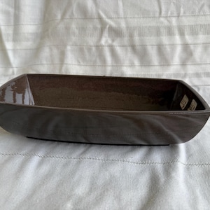 May include: A dark brown rectangular ceramic planter with a boat-like shape. The planter has a glossy interior and a textured exterior with a stitched design along the bottom edge. The planter is suitable for succulents or small plants.
