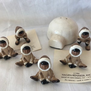 Op de afbeelding: Set van zes keramische beeldjes van mensen in bruine en witte winterkleding, zittend en staand. De beeldjes maken deel uit van een set die een keramisch iglo bevat. De set is gemaakt door Hagen-Renaker, Inc. en is gemaakt in San Dimas, CA, USA. De tekst op de kaart luidt "©1986 Hagen-Renaker, Inc. Gemaakt in San Dimas, CA - U.S.A. (Om te verwijderen - week in water)"