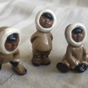 Op de afbeelding: Drie kleine keramische beeldjes van kinderen die bruine en witte winterkleding dragen. De beeldjes staan en zitten.