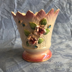 May include: A white ceramic vase with a pink rim and a floral design. The vase features three pink roses with green leaves and blue accents.