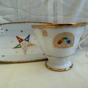 May include: A white porcelain teacup and saucer set with gold trim. The saucer has a five-pointed star with a Masonic symbol in the center. The teacup has a gold trim and a Masonic symbol with a rainbow design.