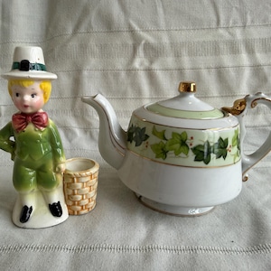 May include: A ceramic figurine of a boy in a green suit, white hat, and red bow tie stands next to a small basket and a white teapot. The teapot has a green and gold ivy leaf design and a gold handle.