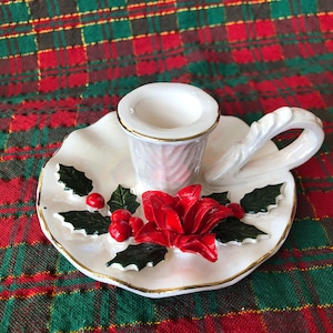 Pode incluir: Castiçal de cerâmica branco com um design de poinsétia e azevinho. O castiçal tem uma alça e uma borda festonada.